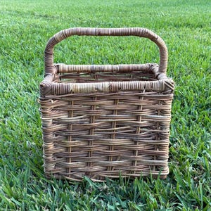May include: A square-shaped woven basket crafted from natural brown wicker material. The basket features a sturdy, arched handle attached to the top rim, with a visible grid-like weave pattern throughout the sides and a flat base for stability.