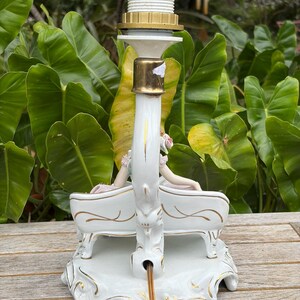 May include: A white ceramic lamp base with gold accents, featuring a swan design with two figures seated on a bench. The lamp has a brass-colored socket and a white lamp holder.