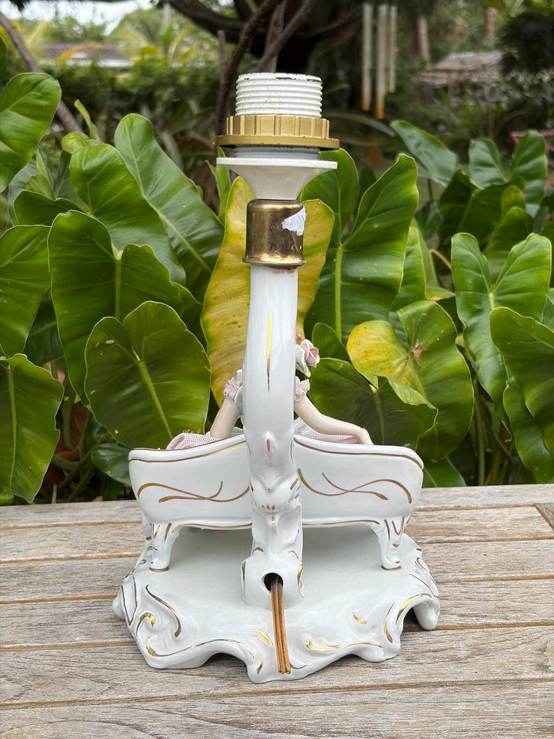 May include: A white ceramic lamp base with gold accents, featuring a swan design with two figures seated on a bench. The lamp has a brass-colored socket and a white lamp holder.