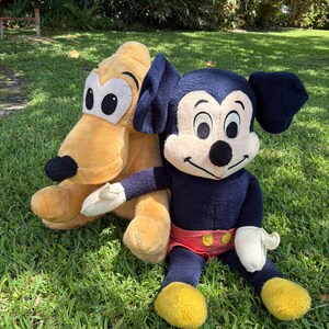May include: Two plush toys of Disney characters, Mickey Mouse and Pluto, are sitting on green grass. Mickey Mouse is wearing a dark blue body, red shorts, yellow shoes, and white gloves. Pluto is a light yellow color.