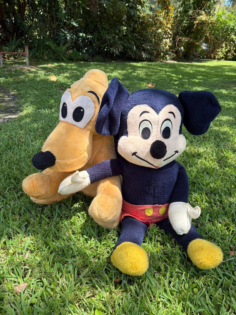 May include: Two plush toys of Disney characters, Mickey Mouse and Pluto, are sitting on green grass. Mickey Mouse is wearing a dark blue body, red shorts, yellow shoes, and white gloves. Pluto is a light yellow color.