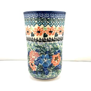 May include: A ceramic tumbler featuring a hand-painted floral pattern with orange and blue blossoms, green leaves, and intricate geometric borders in dark blue and black. The cylindrical vessel has a tapered shape and a smooth, glazed finish.
