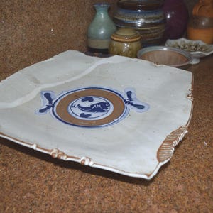 May include: A square, off-white ceramic plate with a brown and blue fish design in the center. The plate has a decorative border and is surrounded by other ceramic items.