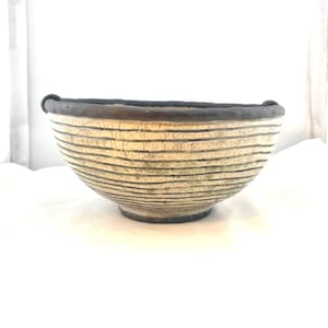May include: A handcrafted ceramic bowl with a cream-colored exterior and horizontal black lines. The rim and base are dark brown. The bowl has a rounded shape and is suitable for serving food or as a decorative piece.