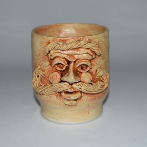 May include: A ceramic mug with a brown and tan face sculpted on the front. The face has a mustache, eyebrows, and a beard.