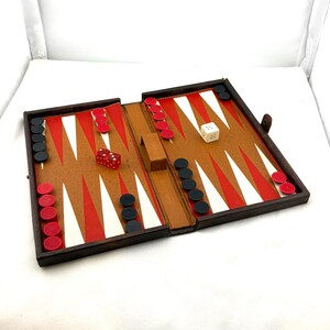 May include: A vintage backgammon set, open and ready to play. The game board features a brown leather-like surface with red and white triangles. Red and black game pieces are arranged on the board, along with two dice. The set is enclosed in a dark brown case.