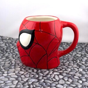 May include: A red ceramic mug shaped like Spider-Man's mask. The mug features a white eye with a black outline, and black web lines across the red surface. The handle is red and curved.