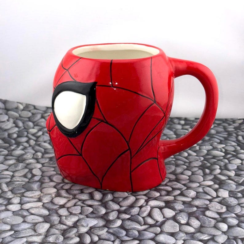 May include: A red ceramic mug shaped like Spider-Man's mask. The mug features a white eye with a black outline, and black web lines across the red surface. The handle is red and curved.
