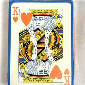 Puede incluir: Una baraja de cartas con el Rey de Corazones. El dise&ntilde;o de la carta incluye una corona amarilla, una ilustraci&oacute;n detallada de un rey y el s&iacute;mbolo del coraz&oacute;n. La baraja tiene un borde azul.