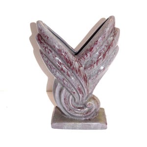 May include: A decorative vase with a unique wing-like design. The vase features a gray base and body with red accents, and a black interior. The base is square, and the body has a swirling, textured pattern.