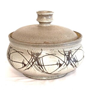 May include: A round ceramic lidded bowl with a speckled beige and off-white finish. The exterior features a dark brown hand-painted abstract pattern of intersecting lines and circular motifs. The lid has a central knob handle for easy lifting.