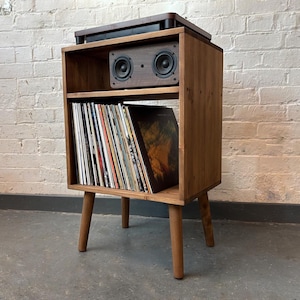 May include: A wooden record player stand with a shelf for vinyl records and a speaker system on top. The stand has four legs and is made of dark brown wood.
