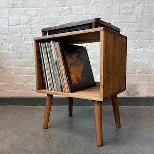 May include: A wooden record player stand with a shelf for storing vinyl records. The stand has a light brown finish and four legs. A record player is sitting on top of the stand.