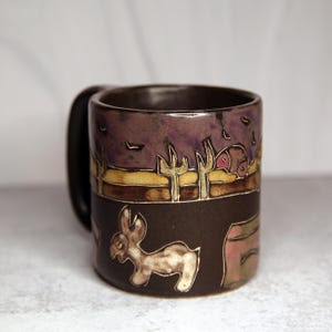 May include: A ceramic mug with a desert landscape design. The mug features a dark brown base with a light-colored donkey illustration. Above, a desert scene with cacti, a purple sky, and a yellow horizon is depicted. The mug has a black handle.