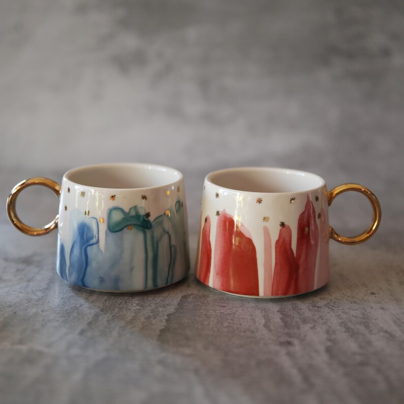 Może przedstawiać: Dwa ceramiczne kubki ze złotymi uchwytami i gwiazdkami. Jeden kubek ma niebieską i turkusową farbę w stylu akwareli, drugi czerwoną i r&oacute;żową. Oba kubki mają białe wnętrze i złoty brzeg.