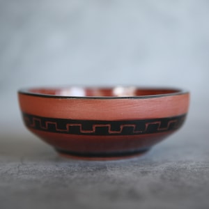 May include: A terracotta-colored ceramic bowl with a black geometric pattern around the middle. The bowl has a simple, rounded shape and a black rim. The background is a soft gray.