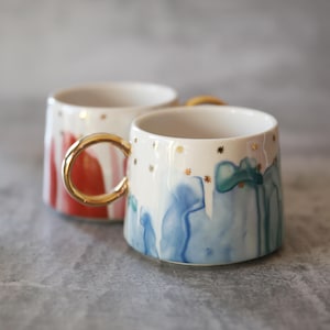 Może przedstawiać: Dwa ceramiczne kubki ze złotymi uchwytami. Jeden kubek ma czerwono-biały wz&oacute;r, drugi niebiesko-zielony wz&oacute;r akwarelowy. Oba kubki mają złote gwiazdki.