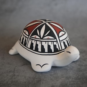 May include: A white ceramic turtle figurine with a painted shell. The shell is decorated with black, red, and white geometric patterns.