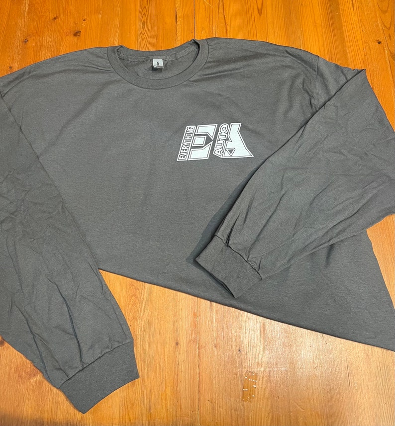 May include: A charcoal gray long-sleeve t-shirt with the text "EVERYTHING AUTO" in a white, blocky font. The shirt has a crew neck and ribbed cuffs. The shirt is made of a soft material.