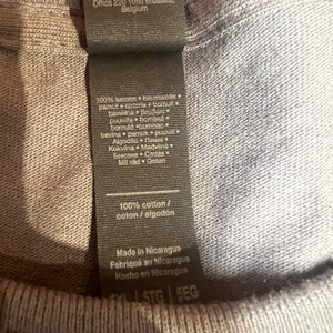 May include: Close-up of a gray t-shirt with a black tag. The tag includes text in multiple languages, stating the shirt is 100% cotton and made in Nicaragua. The shirt's tag also includes the Gildan Activewear EU address.