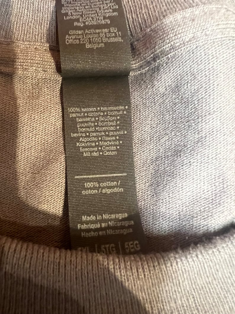 May include: Close-up of a gray t-shirt with a black tag. The tag includes text in multiple languages, stating the shirt is 100% cotton and made in Nicaragua. The shirt's tag also includes the Gildan Activewear EU address.
