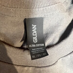 May include: Close-up of a gray t-shirt collar with a black tag. The tag reads "GILDAN ULTRA COTTON" and includes text in English, French, and Spanish. The shirt size is 5XL, 5TG, and 5EG.