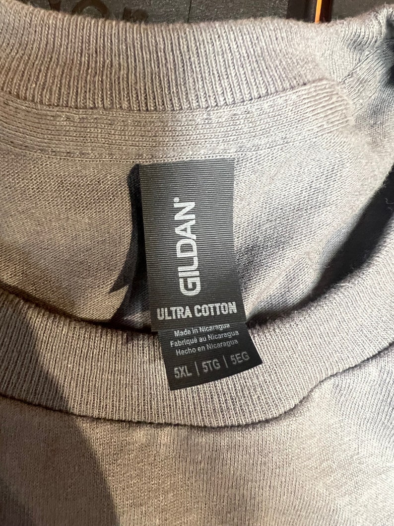 May include: Close-up of a gray t-shirt collar with a black tag. The tag reads "GILDAN ULTRA COTTON" and includes text in English, French, and Spanish. The shirt size is 5XL, 5TG, and 5EG.