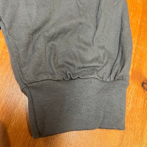 May include: Close-up of a gray long-sleeved shirt cuff. The cuff is ribbed and cinched at the wrist. The fabric of the sleeve is gathered above the cuff. The background is a wooden surface.
