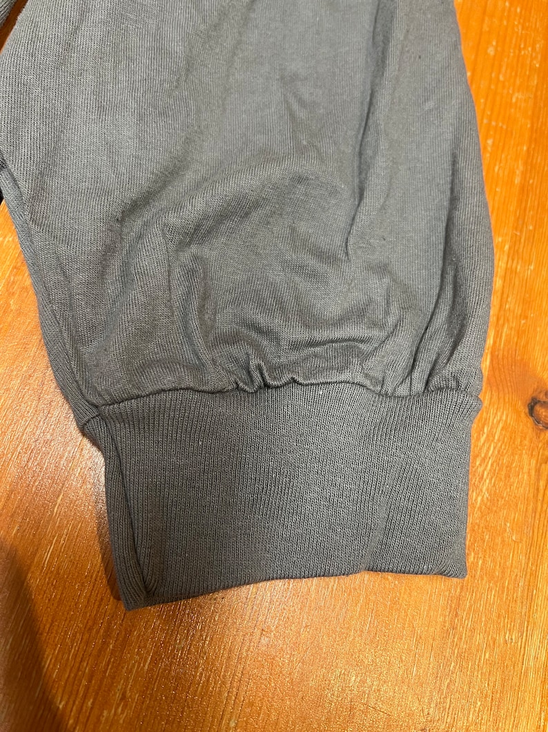 May include: Close-up of a gray long-sleeved shirt cuff. The cuff is ribbed and cinched at the wrist. The fabric of the sleeve is gathered above the cuff. The background is a wooden surface.