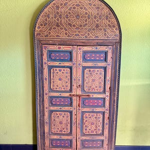 Op de afbeelding: Een gewelfde houten deur met ingewikkelde geometrische patronen in tinten bruin, blauw en rood. De deur heeft een decoratief bovendeel en twee scharnierende panelen met een centrale grendel. De deur staat tegen een lichtgroene muur.