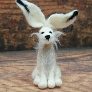 May include: A white felt rabbit with black ears and a long white beard. The rabbit is sitting on a wooden surface.