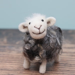May include: A white and gray wool felt sheep with black eyes and a smiling mouth. The sheep is standing on a wooden surface.