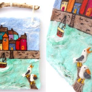 May include: A felt wall hanging depicting a seaside town with colorful houses, a small boat, and two seagulls perched on a wooden post. The background is a blue sky with white clouds and a turquoise sea.