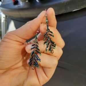May include: A pair of dangle earrings with a leaf design made with black and green beads. The earrings are made with copper wire and have a small bead at the bottom.