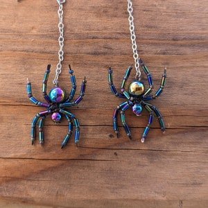 Puede incluir: Un par de pendientes de araña iridiscentes con cadenas plateadas. Las arañas están hechas de cuentas negras y azules con una cuenta redonda iridiscente en el centro.