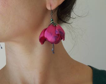 Puede incluir: Primer plano de un pendiente floral rosa y morado. El pendiente está hecho de tela y tiene un gancho de metal oscuro. El pendiente cuelga de la oreja de una persona. El pendiente tiene una pequeña cuenta verde y una cuenta colgante.
