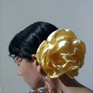 May include: A woman with dark hair is wearing a large, golden, fabric flower in her hair. The flower is made of satin and has a ruffled texture. She is also wearing large, gold, dangling earrings.