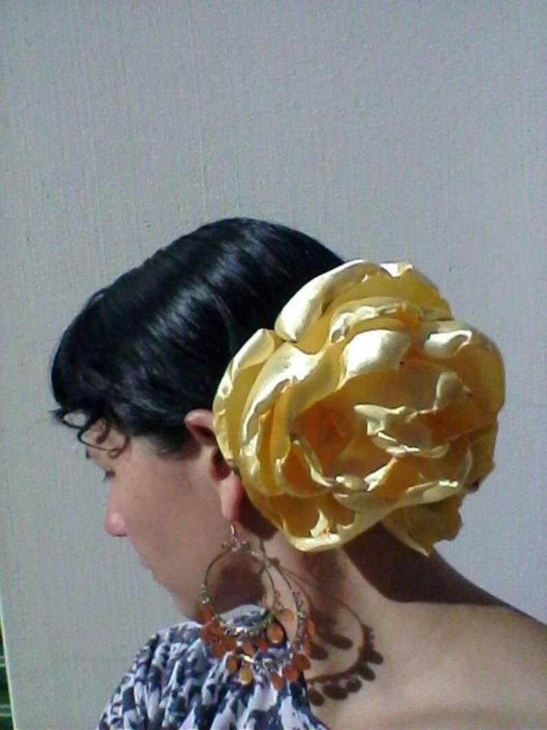 May include: A woman with dark hair is wearing a large, golden, fabric flower in her hair. The flower is made of satin and has a ruffled texture. She is also wearing large, gold, dangling earrings.