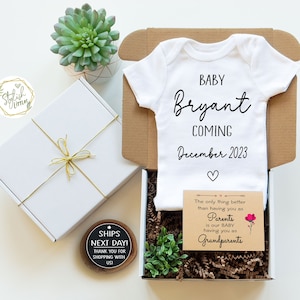 May include: A white baby onesie with the text "BABY Bryant COMING December 2023" in black. The onesie is in a brown cardboard box with a white gift box, a small wooden sign that says "SHIPS NEXT DAY! THANK YOU FOR SHOPPING WITH USI", and a small card that says "The only thing better than having you as Parents is our BABY having you as Grandparents".