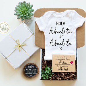 May include: A white baby onesie with black text that reads "HOLA Abuelita y Abuelito" with a small pink heart. The onesie is in a brown cardboard box with a small card that reads "Lo unico mejor que tenerlos como Padres es que nuestro bebe los tenga como Abuelos!" with a pink flower graphic.