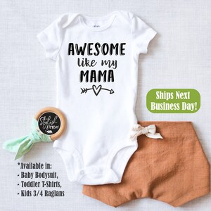 Op de afbeelding: Witte baby-romper met zwarte tekst die "AWESOME like my MAMA" luidt, met een pijl die naar een hart wijst. De romper is op een witte achtergrond.