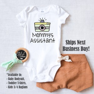 May include: White baby onesie with a cartoon camera and the text "Mommy's Assistant".