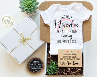 May include: A white onesie with a rainbow design and the text "The Best Miracles Take A Little Time Arriving December 2023". A brown card with the text "Could be pink, could be blue, all we know is that We Are Due!" is in the box with the onesie. The box is filled with brown crinkle paper and a small green plant.