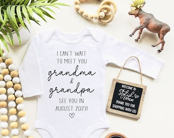 May include: White long-sleeve bodysuit with the text "I can't wait to meet you grandma & grandpa see you in August 2023!"  A small wooden sign with the text "Welcome to Stylish Littles Thank you for shopping with us!" is hanging on the right side of the bodysuit.