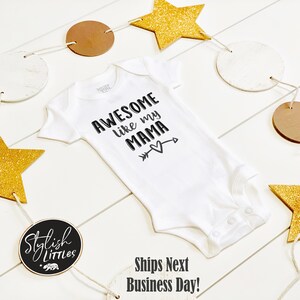 Op de afbeelding: Witte baby romper met zwarte tekst die "Awesome like my mama" luidt, met een pijl die naar een hart wijst. De romper is omringd door een guirlande van gouden sterren en bruine en witte cirkels.