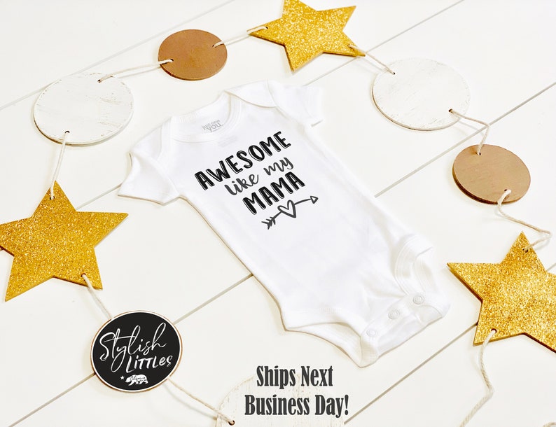 Op de afbeelding: Witte baby romper met zwarte tekst die "Awesome like my mama" luidt, met een pijl die naar een hart wijst. De romper is omringd door een guirlande van gouden sterren en bruine en witte cirkels.