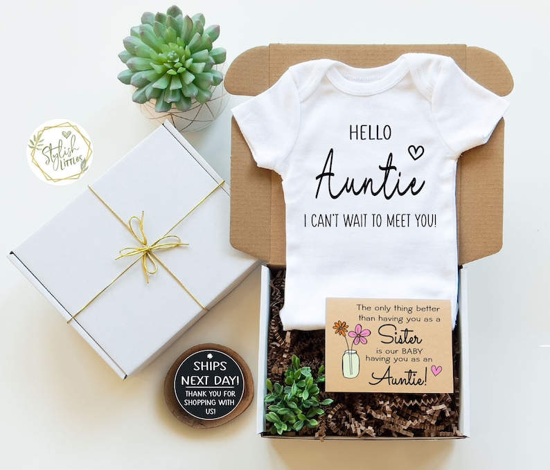 May include: A white baby onesie with the text "Hello Auntie I can't wait to meet you!"  A card with the text "The only thing better than having you as a Sister is our Baby having you as an Auntie!"  The card has a floral design and a heart. The onesie and card are in a white gift box with gold ribbon.