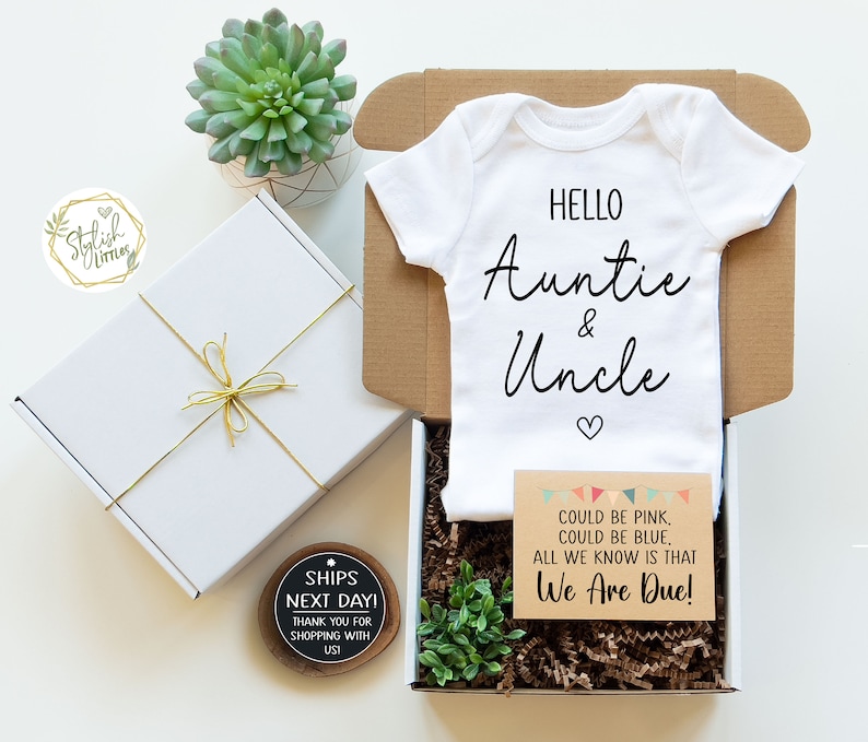 May include: A white baby onesie with the text "Hello Auntie & Uncle" in black script. The onesie is in a gift box with a card that says "Could be pink, could be blue. All we know is that We Are Due!"