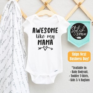 Op de afbeelding: Witte baby romper met zwarte tekst die "Awesome like my Mama" luidt, met een pijl- en hartvormig motief. De romper hangt aan een houten hanger.