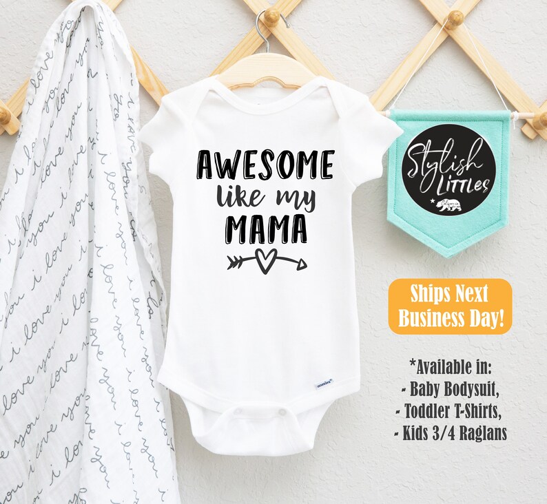 Op de afbeelding: Witte baby romper met zwarte tekst die "Awesome like my Mama" luidt, met een pijl- en hartvormig motief. De romper hangt aan een houten hanger.